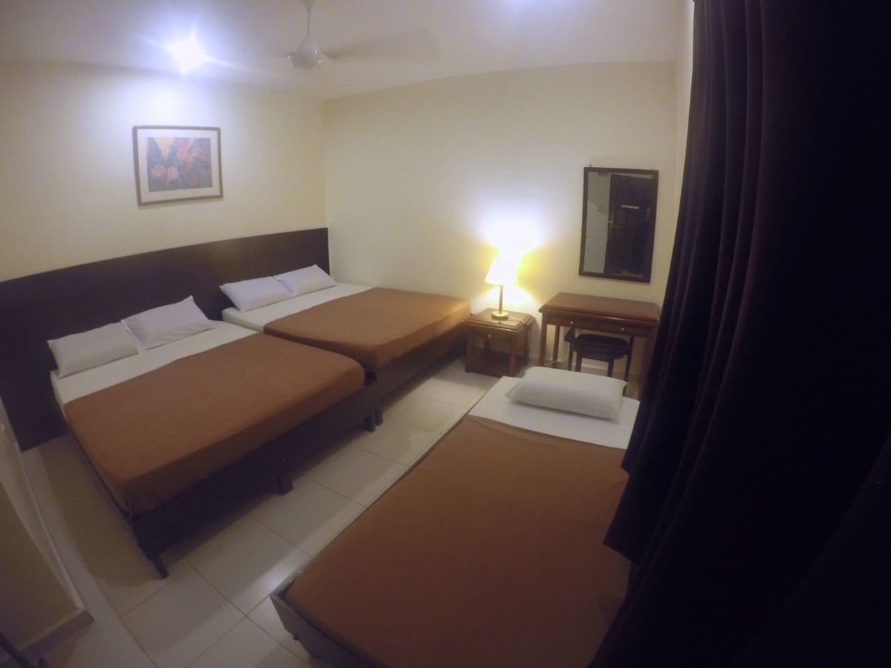 A hotel room with three beds, a bedside table with a lamp, a small table, and a mirror.
