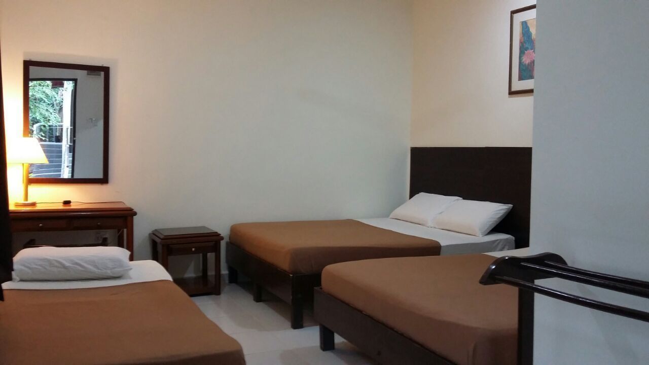 A hotel room with three beds, a bedside table with a lamp, a small table, and a mirror on the wall.