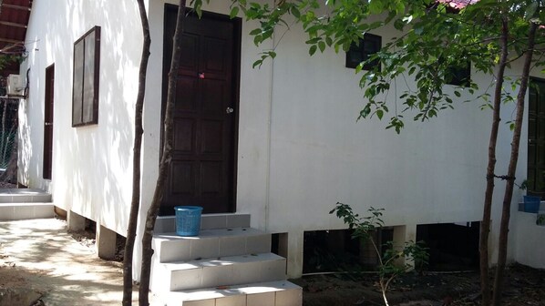 Un edificio blanco con puerta y ventanas marrón oscuro, rodeado de árboles y un pequeño jardín.