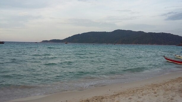 A beach with calm waters, a boat on the shore, and a forested hill in the background. - Perhentian Tropicana Inn (Pulau Perhentian Kecil)