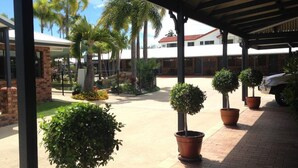 A covered outdoor area with potted plants, a paved walkway, and a building in the background. - Country Ayr Motel (Ayr)