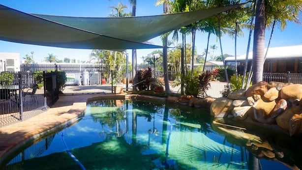 A clear swimming pool with a rock feature, surrounded by palm trees and a shaded area. - Country Ayr Motel (Ayr)