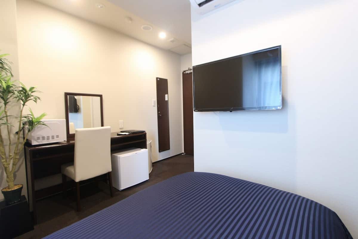 Habitación de hotel con cama, escritorio y silla, refrigerador pequeño, planta en maceta, espejo y televisor montado en la pared.