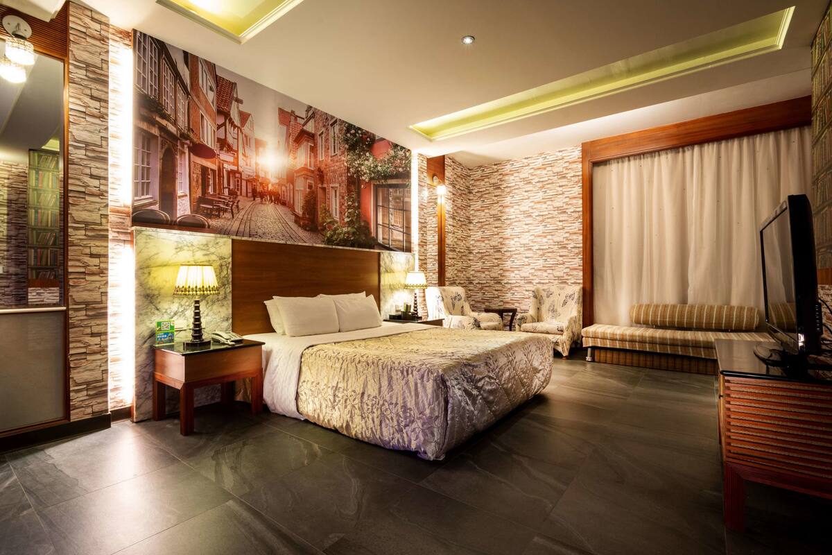 A well-lit hotel room with a large bed, a seating area with a sofa and armchairs, a television, and a bookshelf.