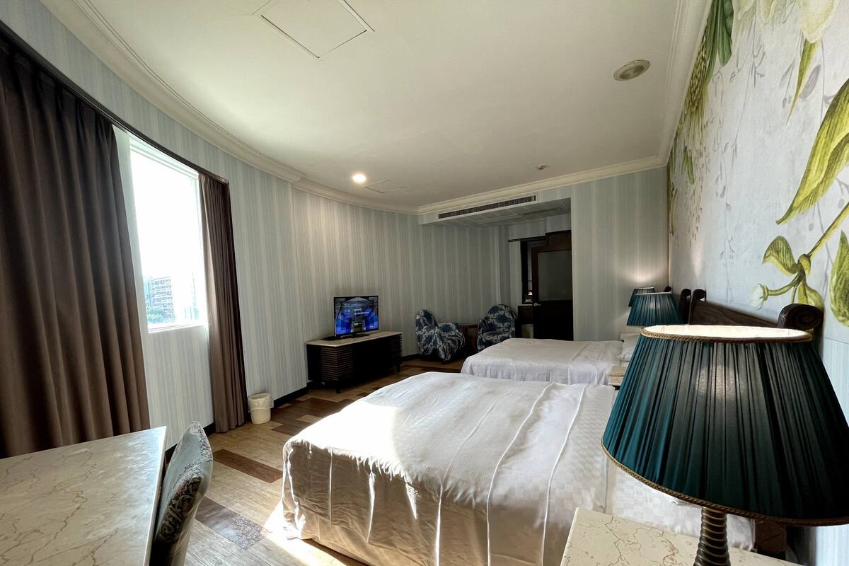 A hotel room with two beds, a TV, a desk, and a large window with curtains.