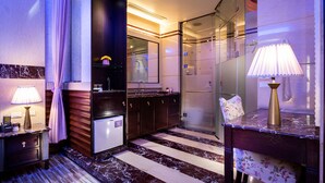 Un baño moderno con encimera de mármol, un refrigerador pequeño, cabina de ducha de vidrio y tocador con espejo y lámpara.