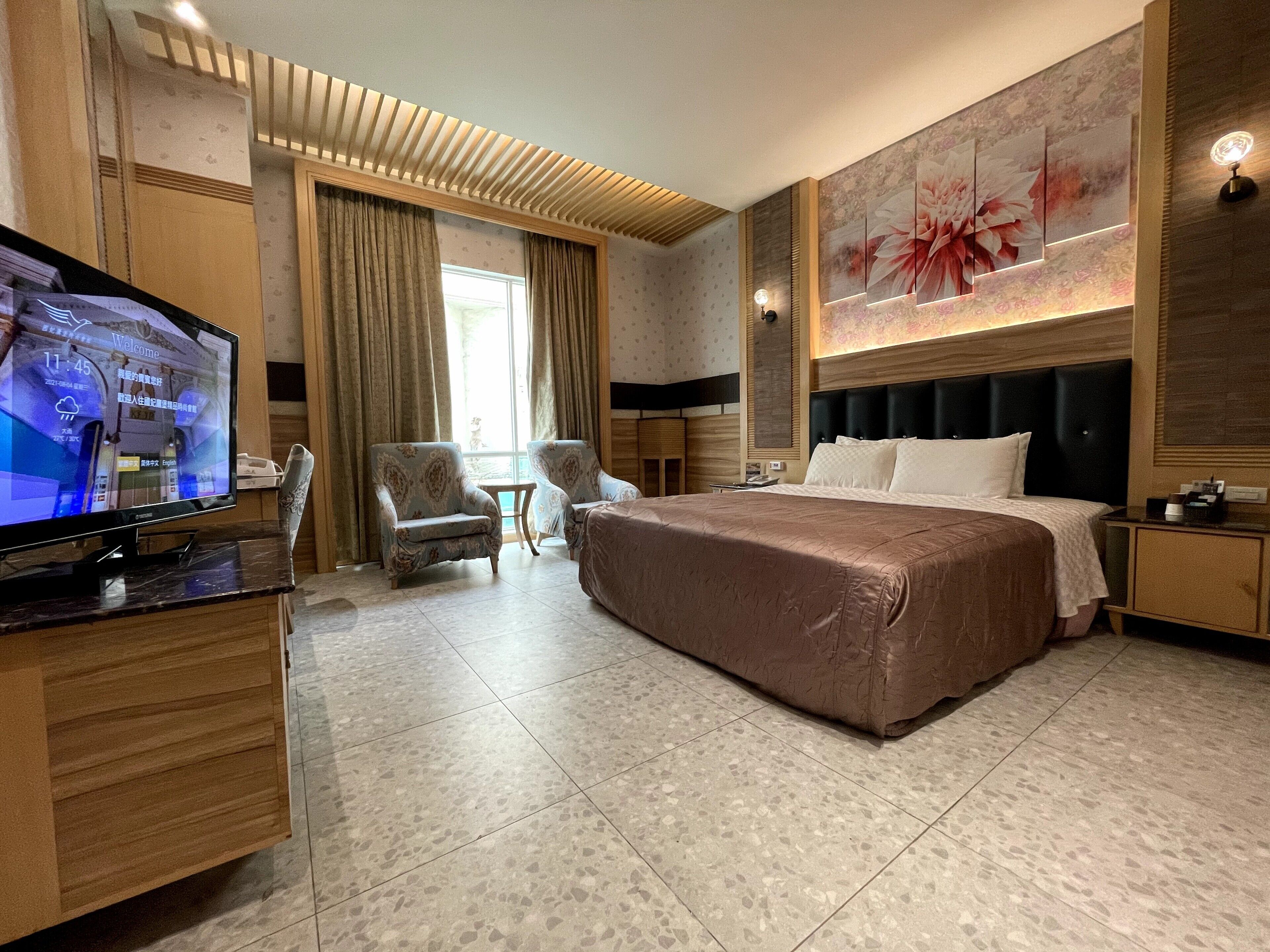 Habitación de hotel con cama, escritorio con televisor, una silla, una mesita y un cuadro floral en la pared.