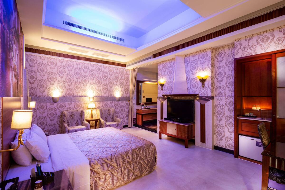 A hotel room with a large bed, a TV, a fireplace, and a desk with a lamp.