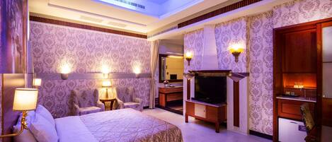 A hotel room with a large bed, a TV, a fireplace, and a desk with a lamp.