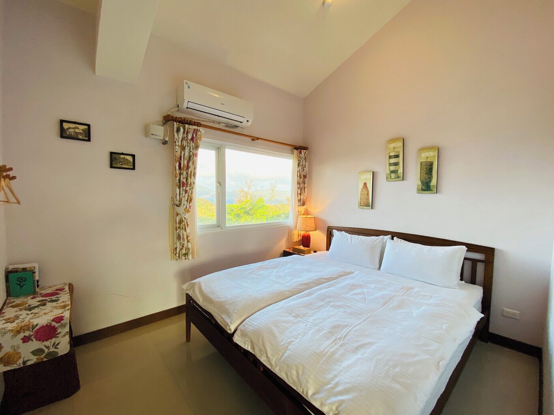 Un dormitorio con cama, aire acondicionado, ventana con cortinas, mesita de noche con lámpara y una obra de arte en la pared.