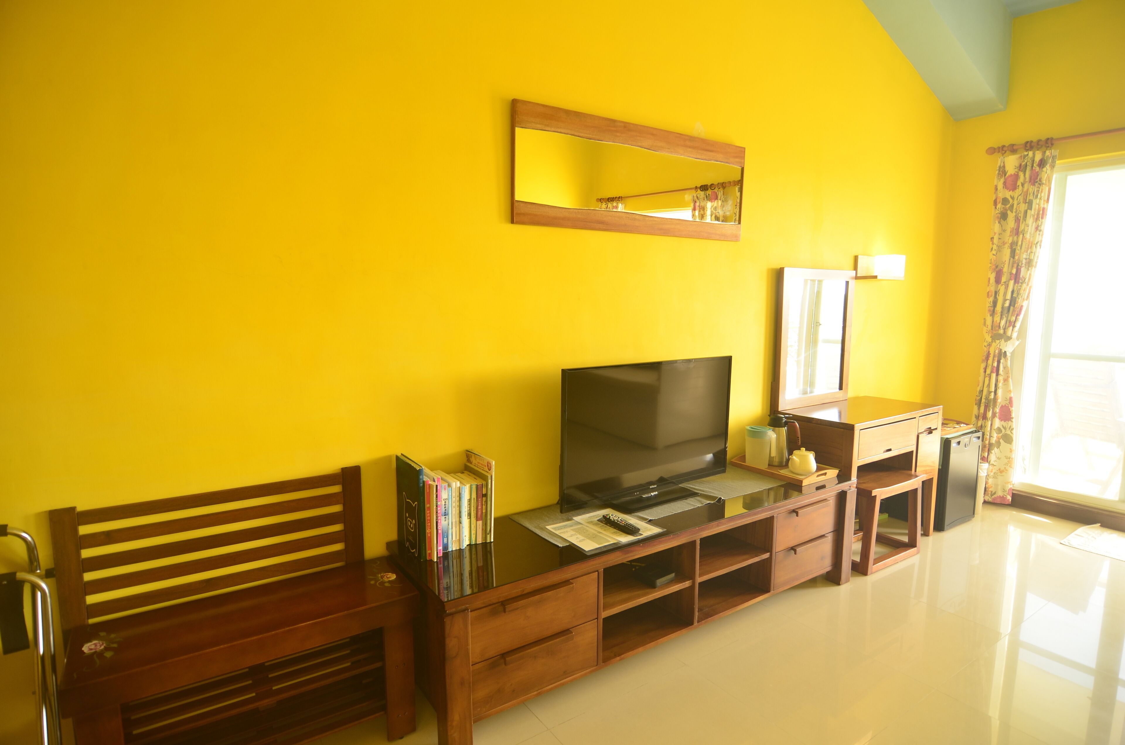 Una habitación con un banco de madera, un televisor de pantalla plana, una pequeña mesa con libros, un espejo y una ventana con cortinas.