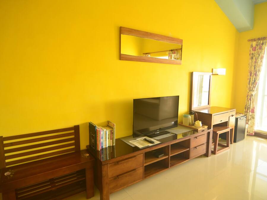 Una habitación con un banco de madera, un televisor de pantalla plana, una pequeña mesa con libros, un espejo y una ventana con cortinas.