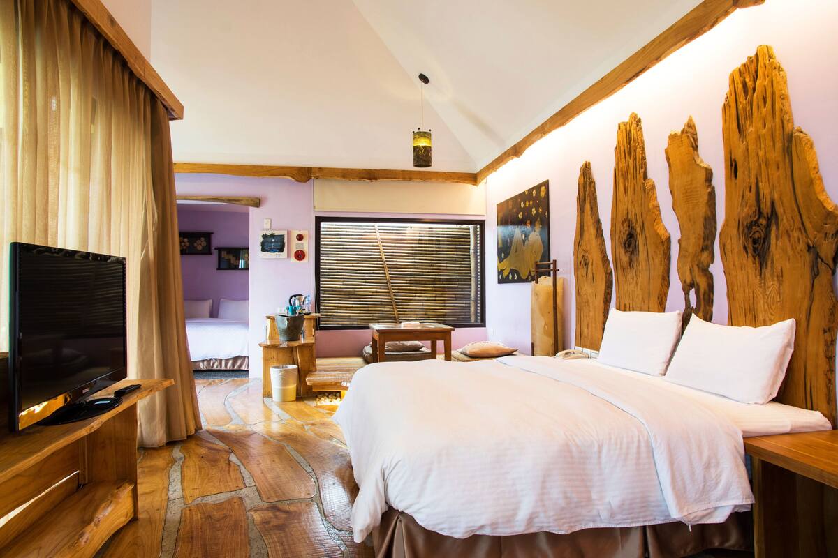 A bedroom with a large bed, a television on a wooden stand, a window with blinds, and a wooden headboard.