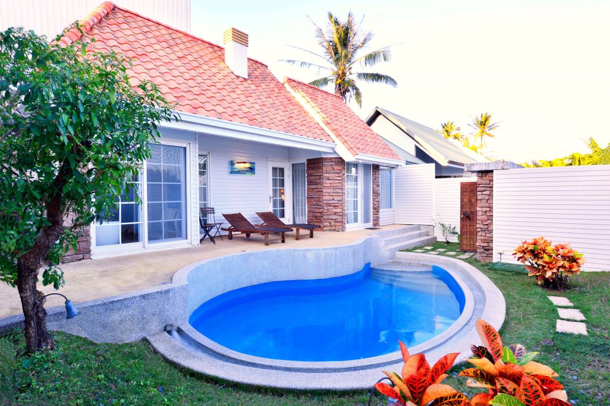 A house with a swimming pool, a patio with chairs, and a garden with plants.