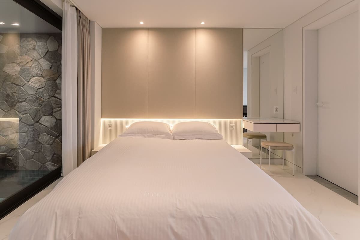 A modern bedroom with a stone accent wall, a large bed, a bedside mirror, and a glass-enclosed shower area.