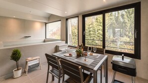 Un comedor con una mesa preparada para dos personas, una pequeña planta en maceta y vistas a árboles a través de amplios ventanales.