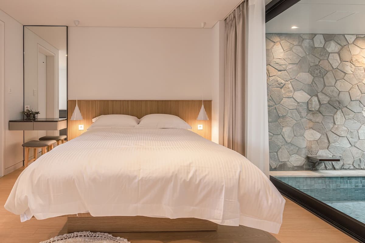 A modern bedroom with a large bed, wooden headboard, bedside lamps, and a stone wall.