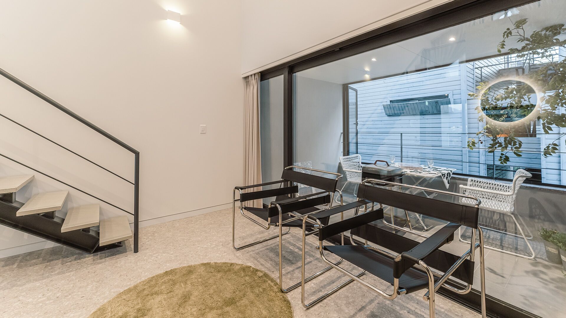 Een modern interieur met een trap, een eethoek met stoelen met metalen frame, en een groot raam met uitzicht op een gebouw en groen.