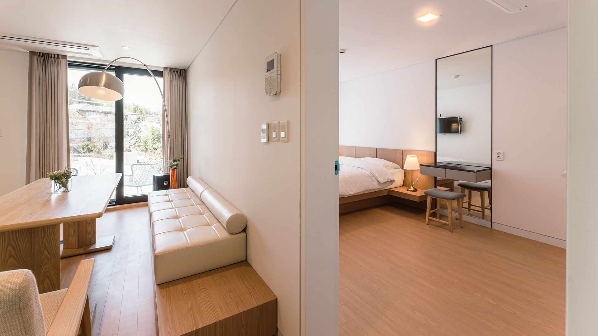 A modern hotel room with a wooden desk, a cushioned bench, a bed with a nightstand, a lamp, and a large mirror.