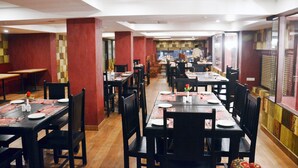 Un restaurante con varias mesas y sillas, dispuesto en un espacio amplio con techos altos y paredes combinadas en tonos rojo y beige.