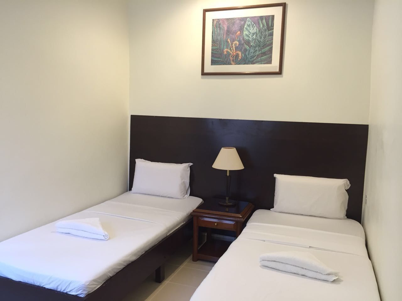 A hotel room with two single beds, a nightstand with a lamp, and a framed picture on the wall.