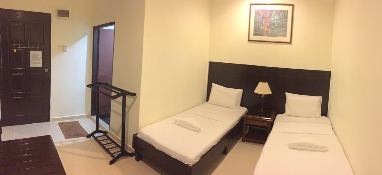 Habitación de hotel con dos camas individuales, una mesita de noche con lámpara, un banco de madera, una puerta y un cuadro en la pared.