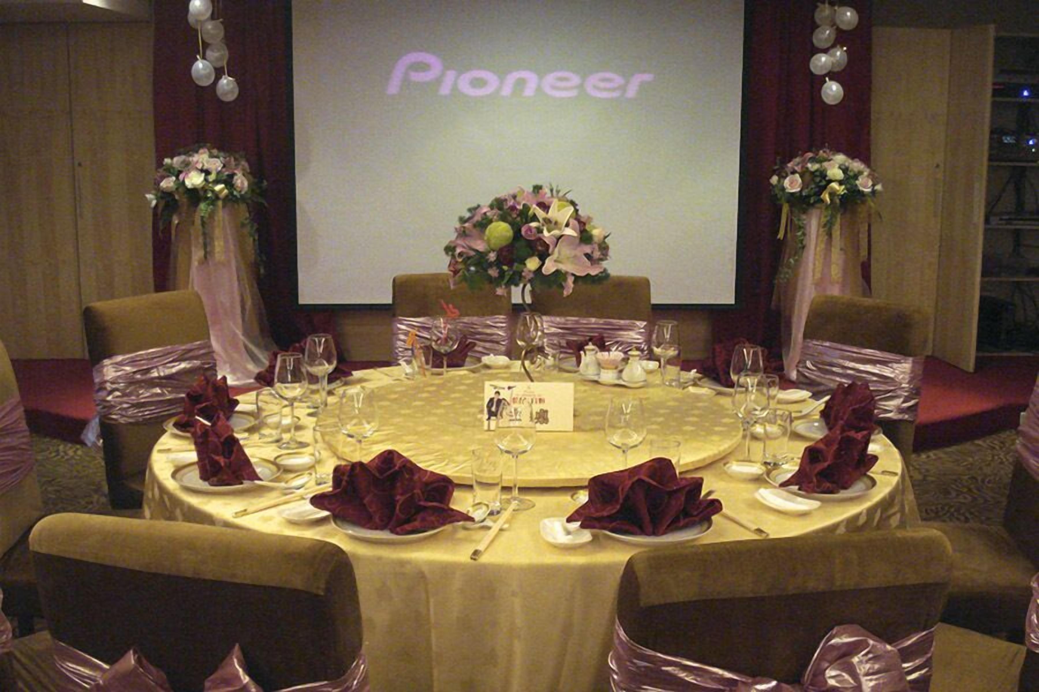 A conference room with round tables set for a formal event, a large screen displaying the word "Pioneer," and floral centerpieces.