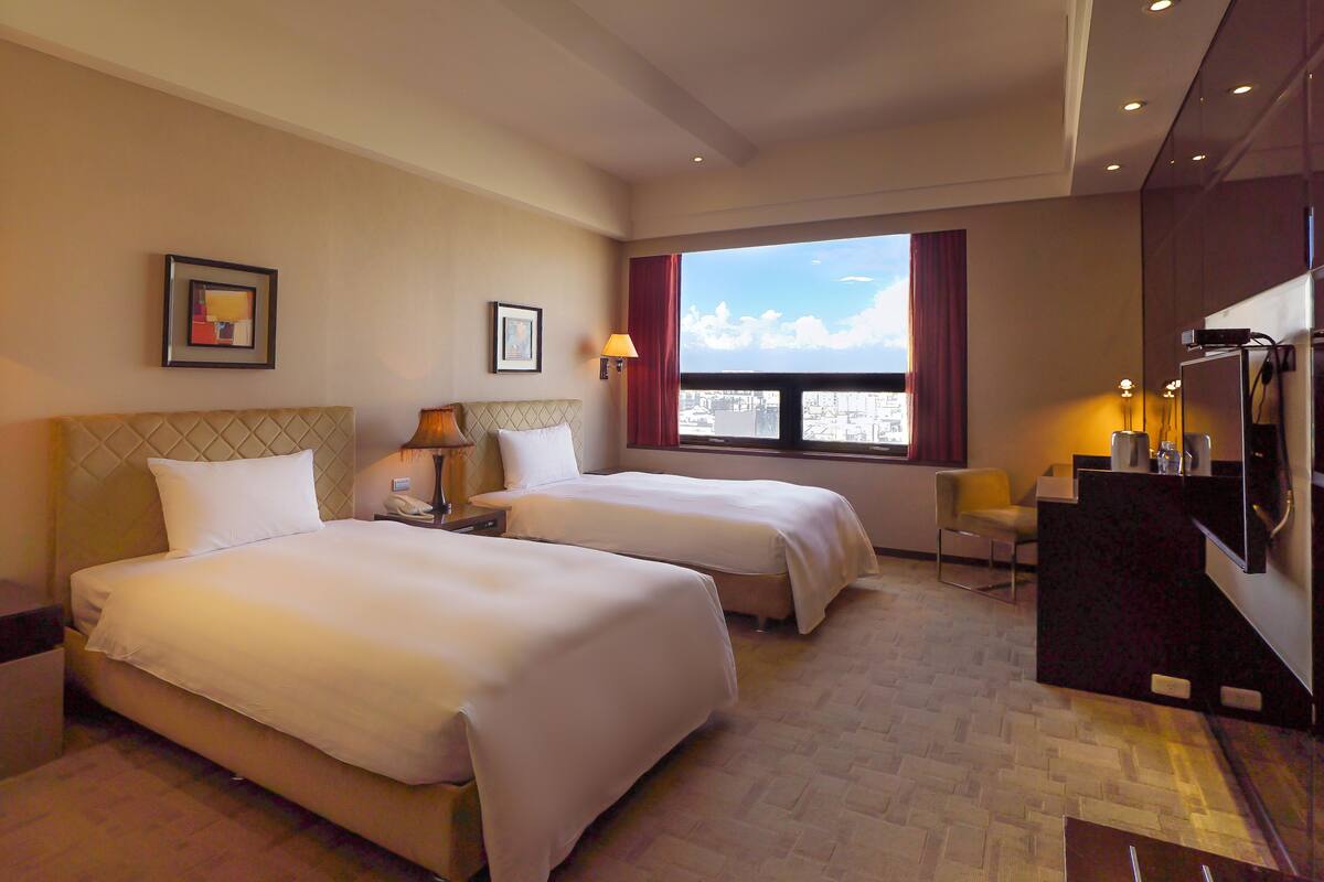 Habitación de hotel con dos camas, un escritorio, una silla, un televisor y una ventana con vista a la ciudad.