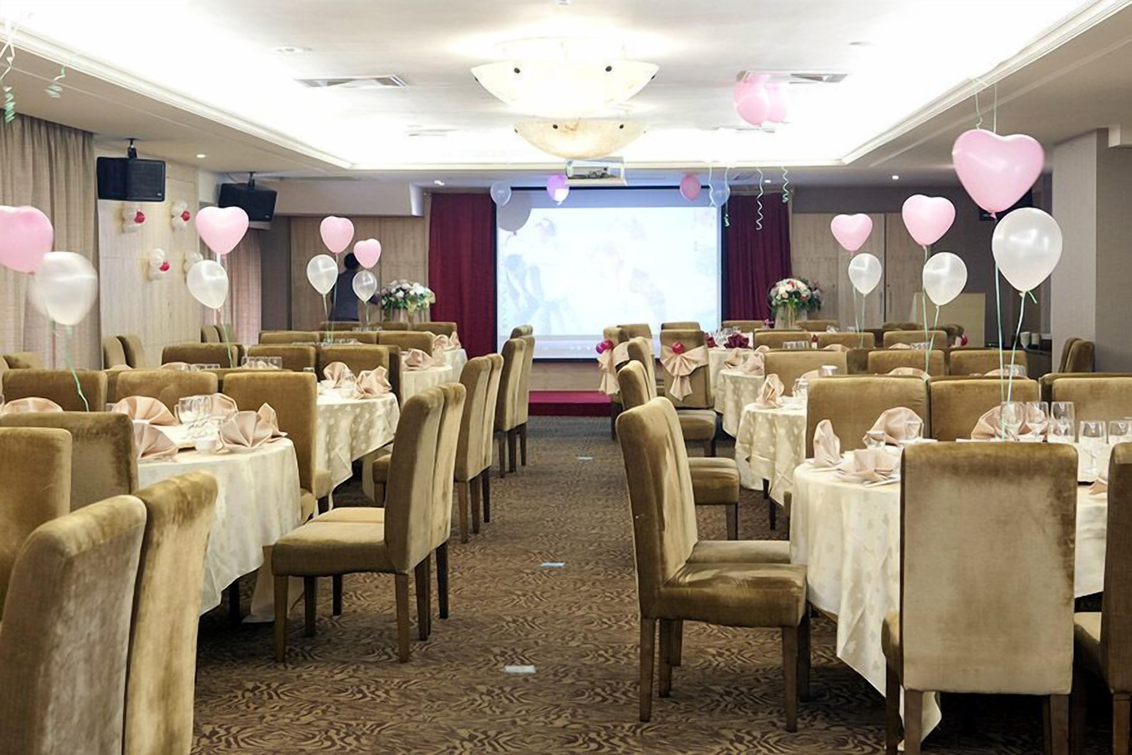 A banquet hall with rows of tables and chairs, a stage with a screen, and balloons.
