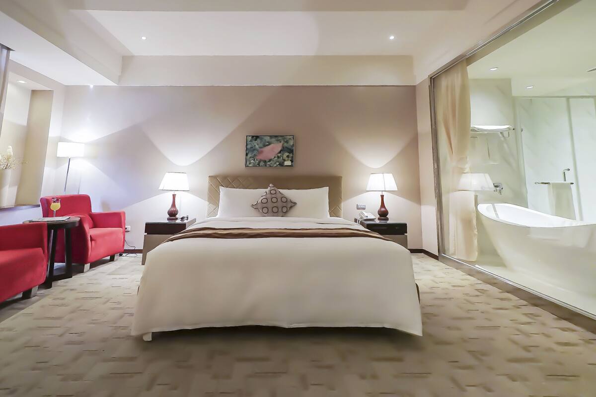Habitación de hotel con una cama grande, dos mesitas de noche con lámparas, un sillón rojo, un espejo grande y vista a un baño con bañera.