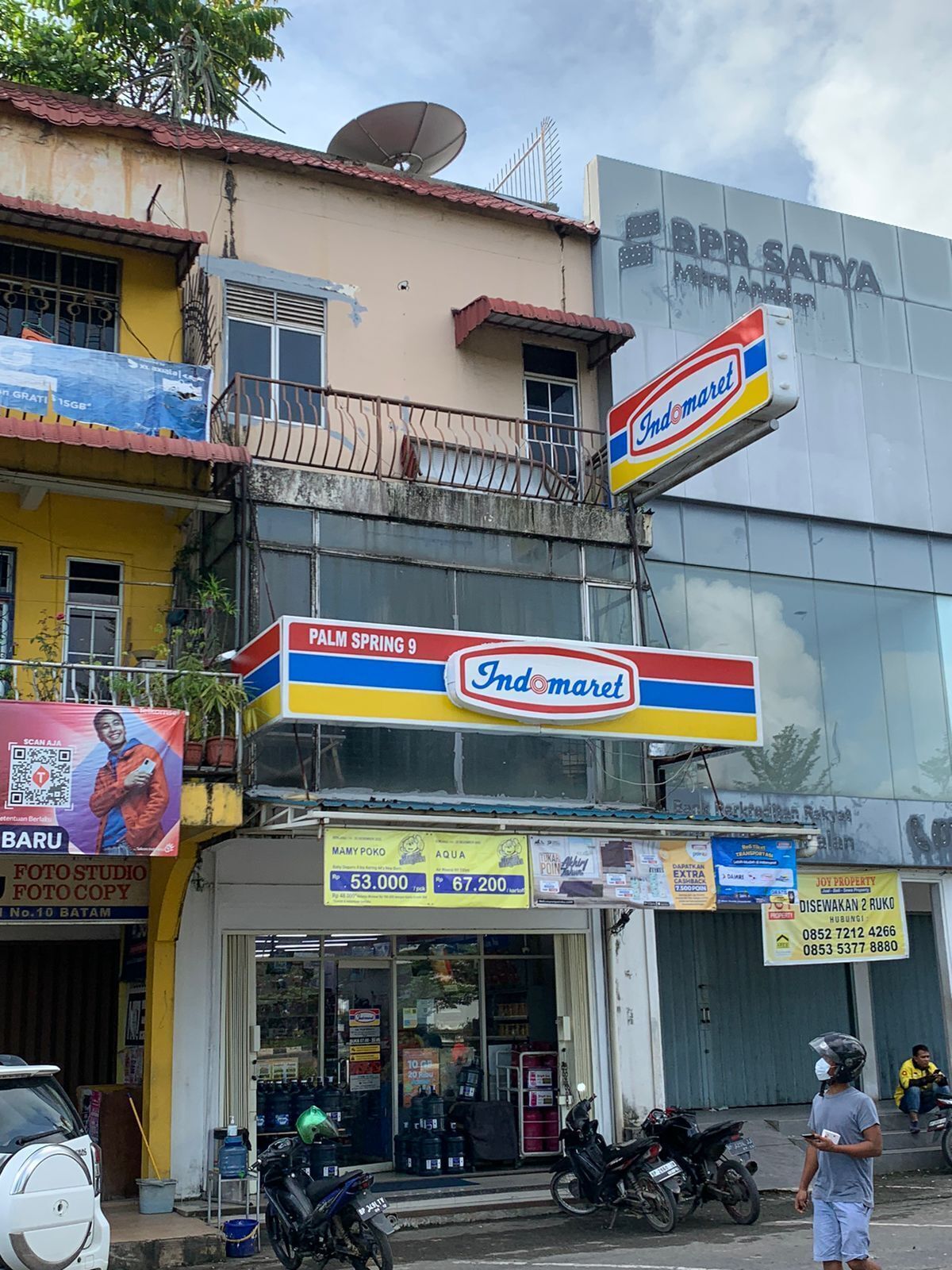 Un escaparate con un letrero de Indomaret, una motocicleta estacionada al frente y un edificio con una antena parabólica.