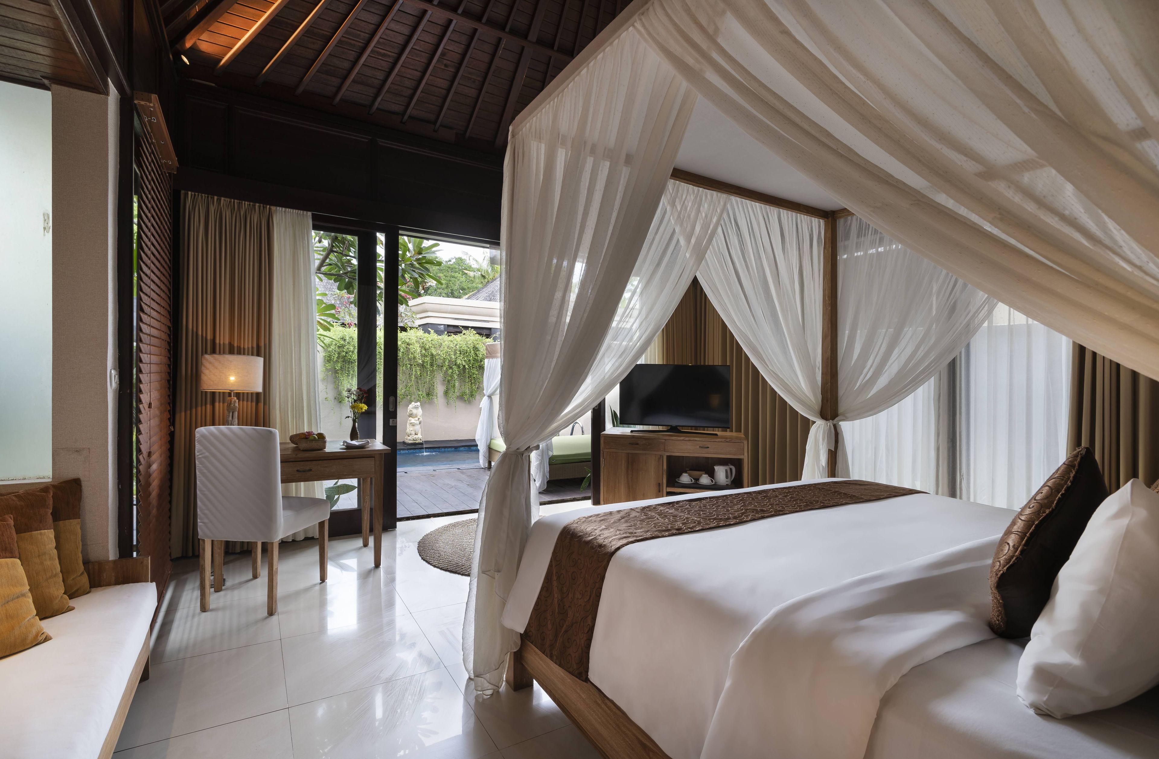 A bedroom with a canopy bed, a desk with a chair, a television, and a view of the outdoors.