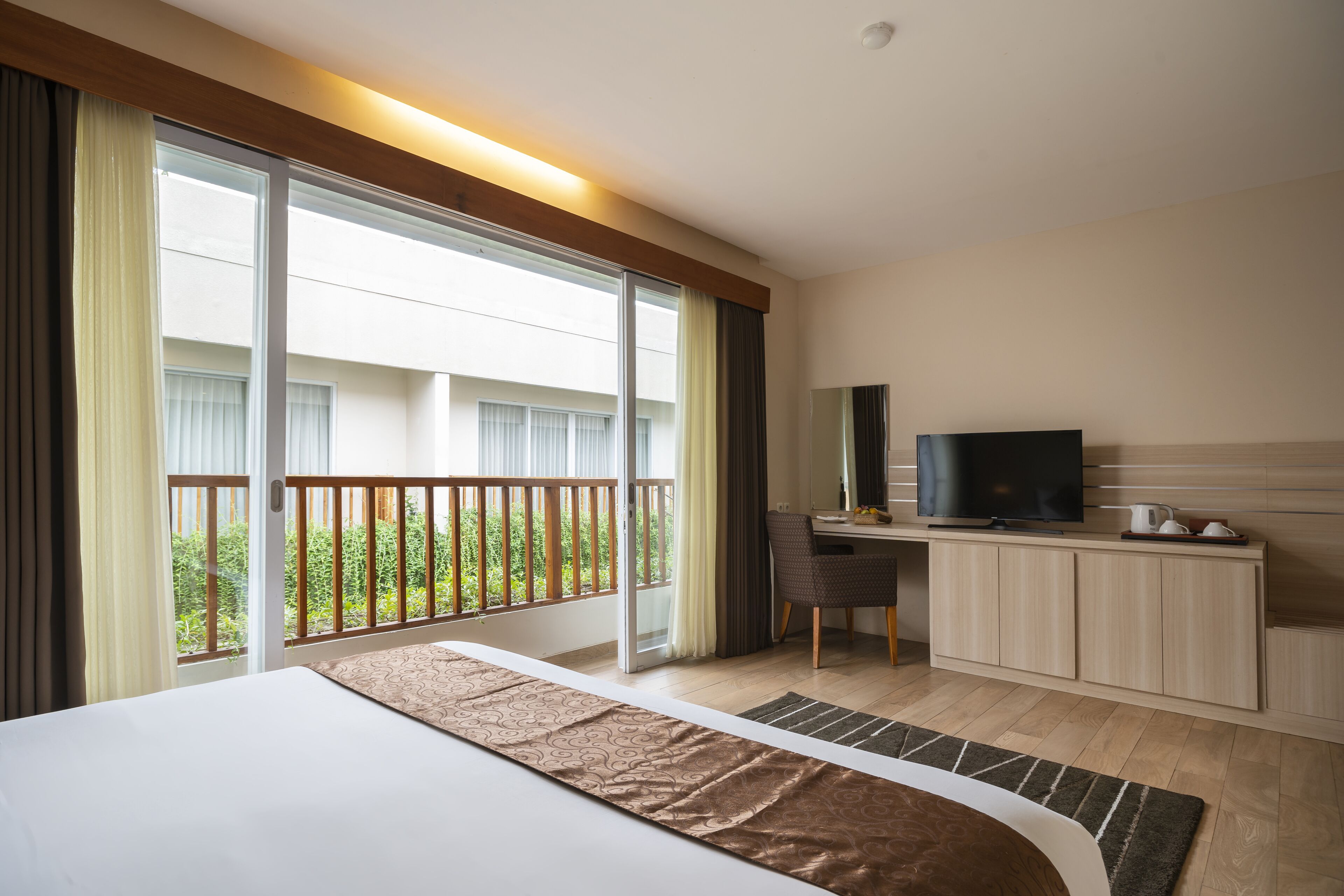 A hotel room with a large bed, a desk with a television, and a balcony with a view of greenery.