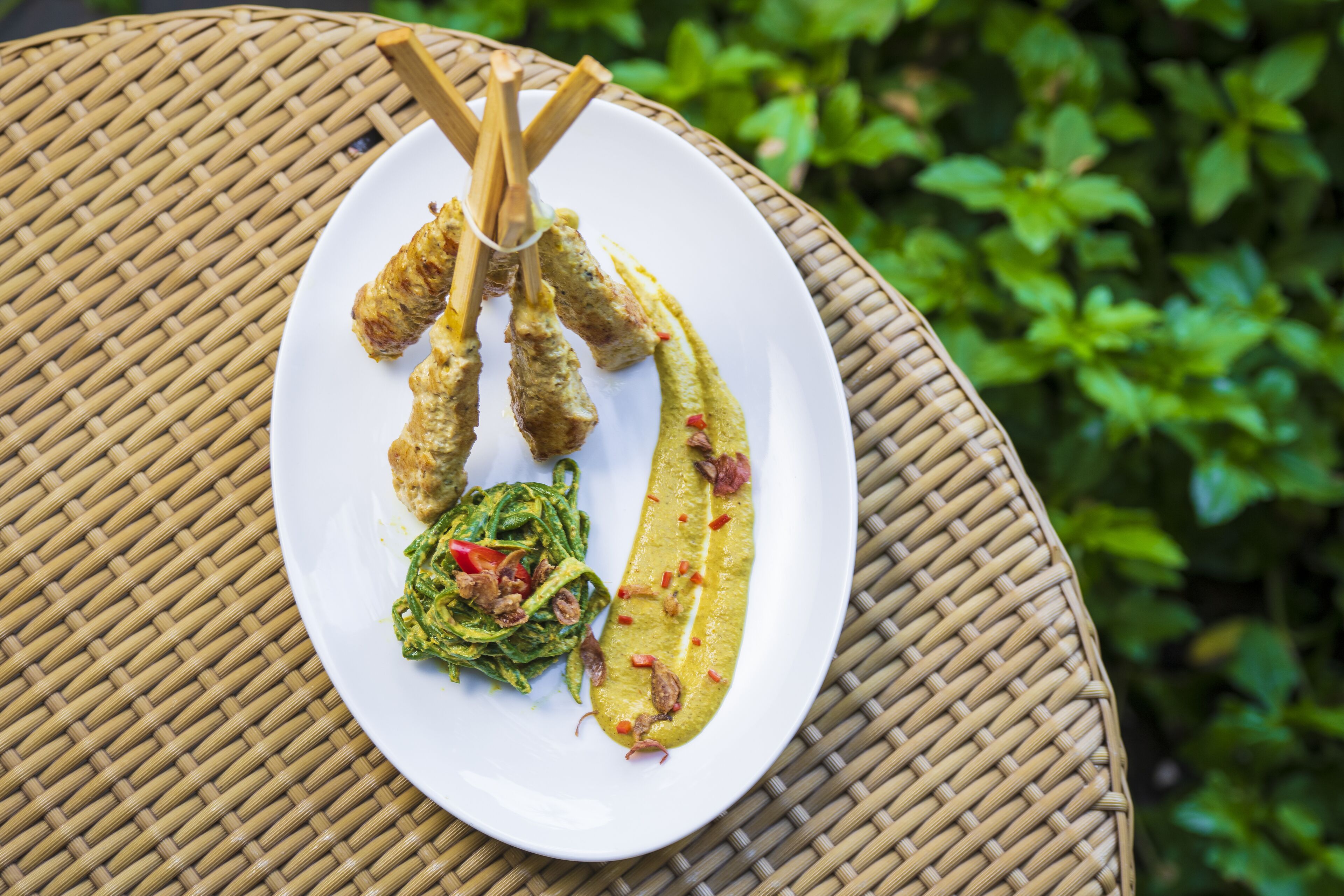 A plate with grilled skewers, a long sausage, and a side of greens on a woven table.