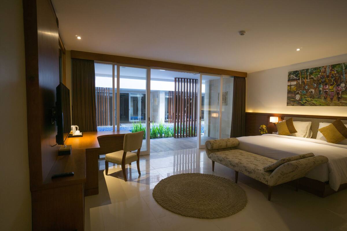 A modern hotel room with a large bed, a desk with a chair, a television, and a view of the outdoors through a sliding glass door.