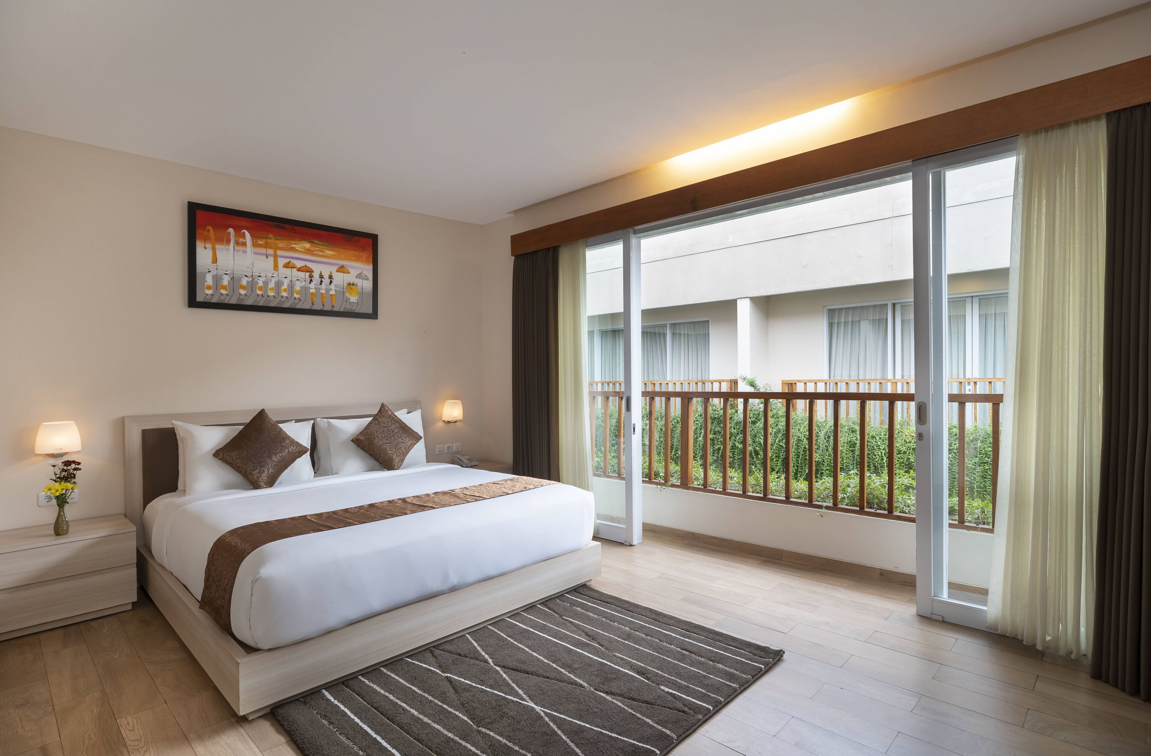 A hotel room with a large bed, a nightstand, a lamp, a painting, and a balcony with a view of greenery.