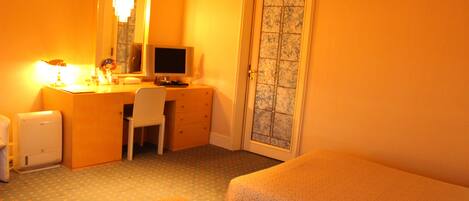 A hotel room with a bed, a desk with a computer, a chair, and a small table with a lamp.