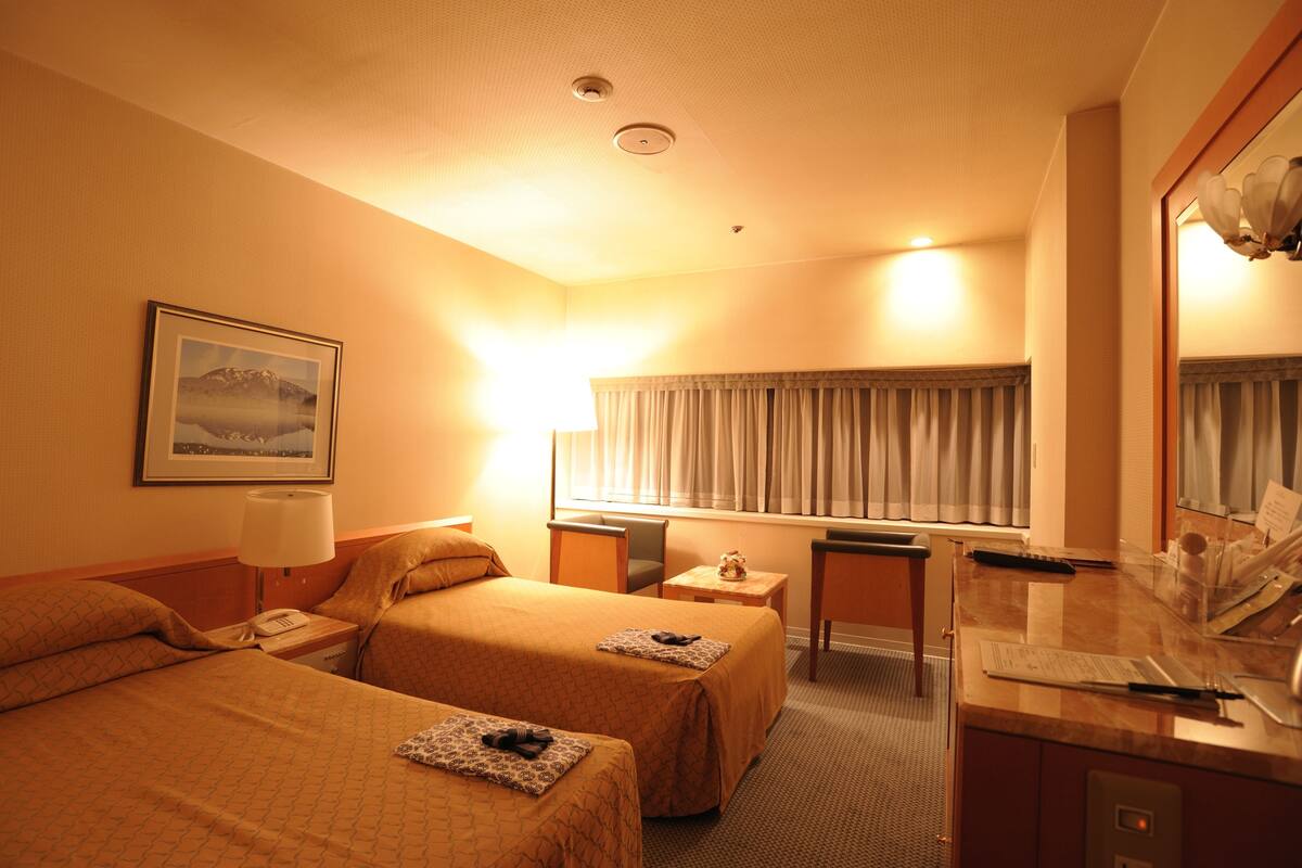 A hotel room with two beds, a desk, a chair, a lamp, a framed picture on the wall, and a window with curtains.