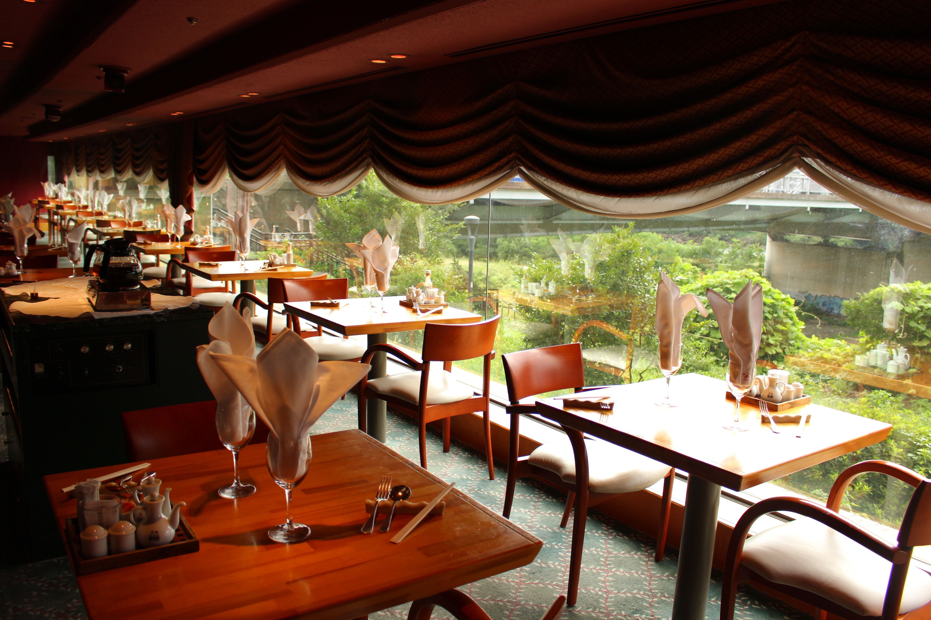 A restaurant with tables set for dining, featuring a view of greenery through large windows.