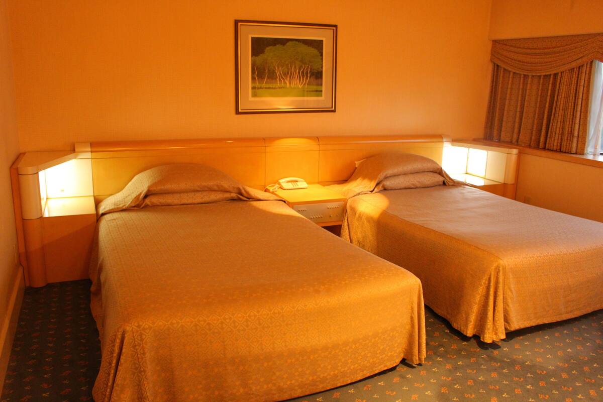 A hotel room with two single beds, a bedside table with a lamp, a framed picture on the wall, and a window with curtains.