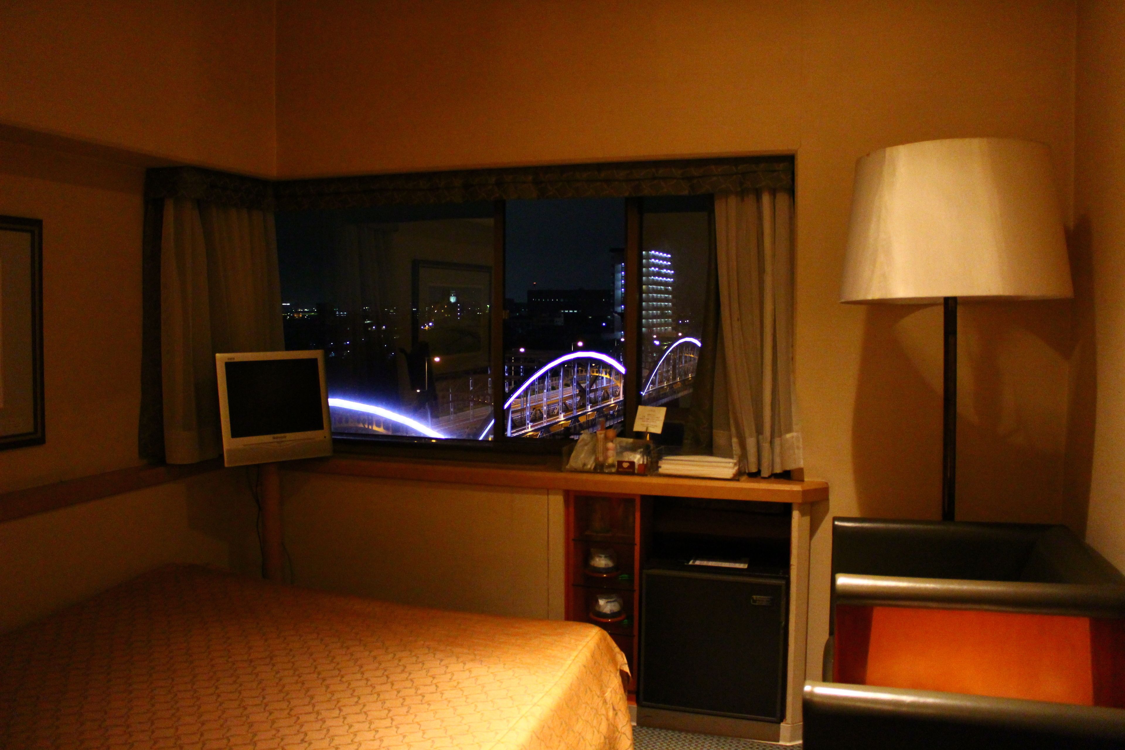 A hotel room with a bed, a desk with a television, a lamp, and a view of a cityscape with a bridge at night.