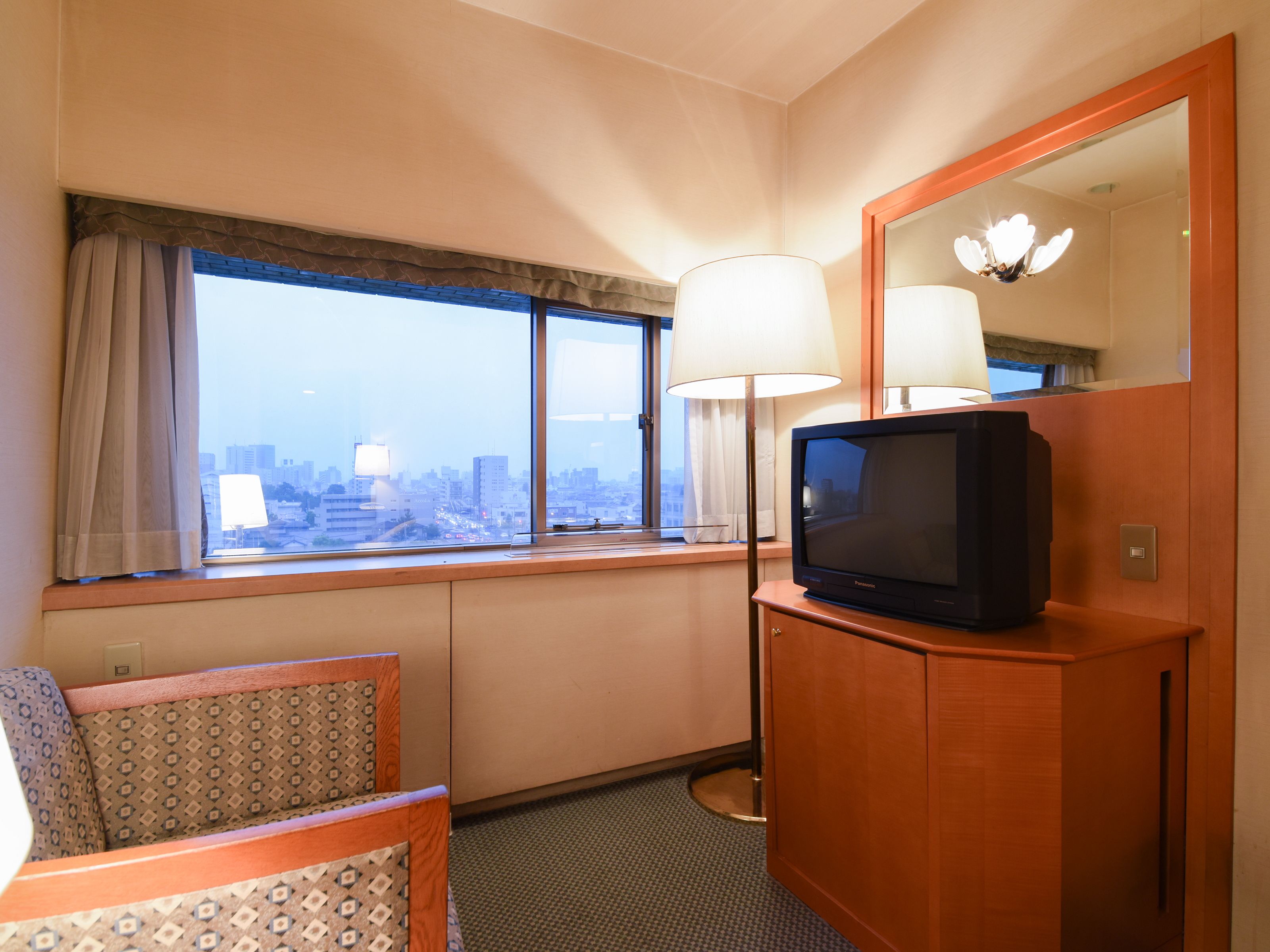 A hotel room with a large window, a television on a wooden cabinet, a lamp, and a patterned chair.