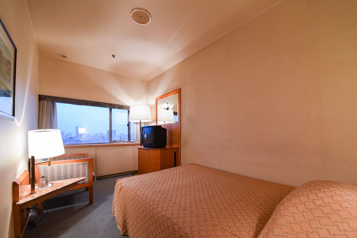 A hotel room with a bed, a desk with a lamp, a television, and a window with a city view.
