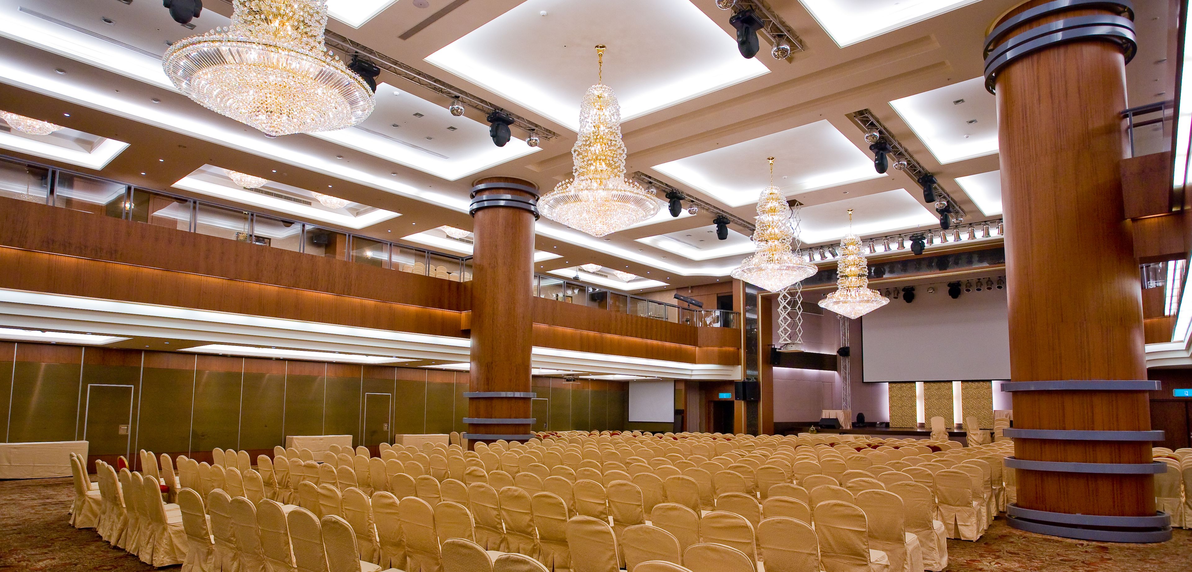 A large conference room with rows of chairs, chandeliers, and a stage with a screen.