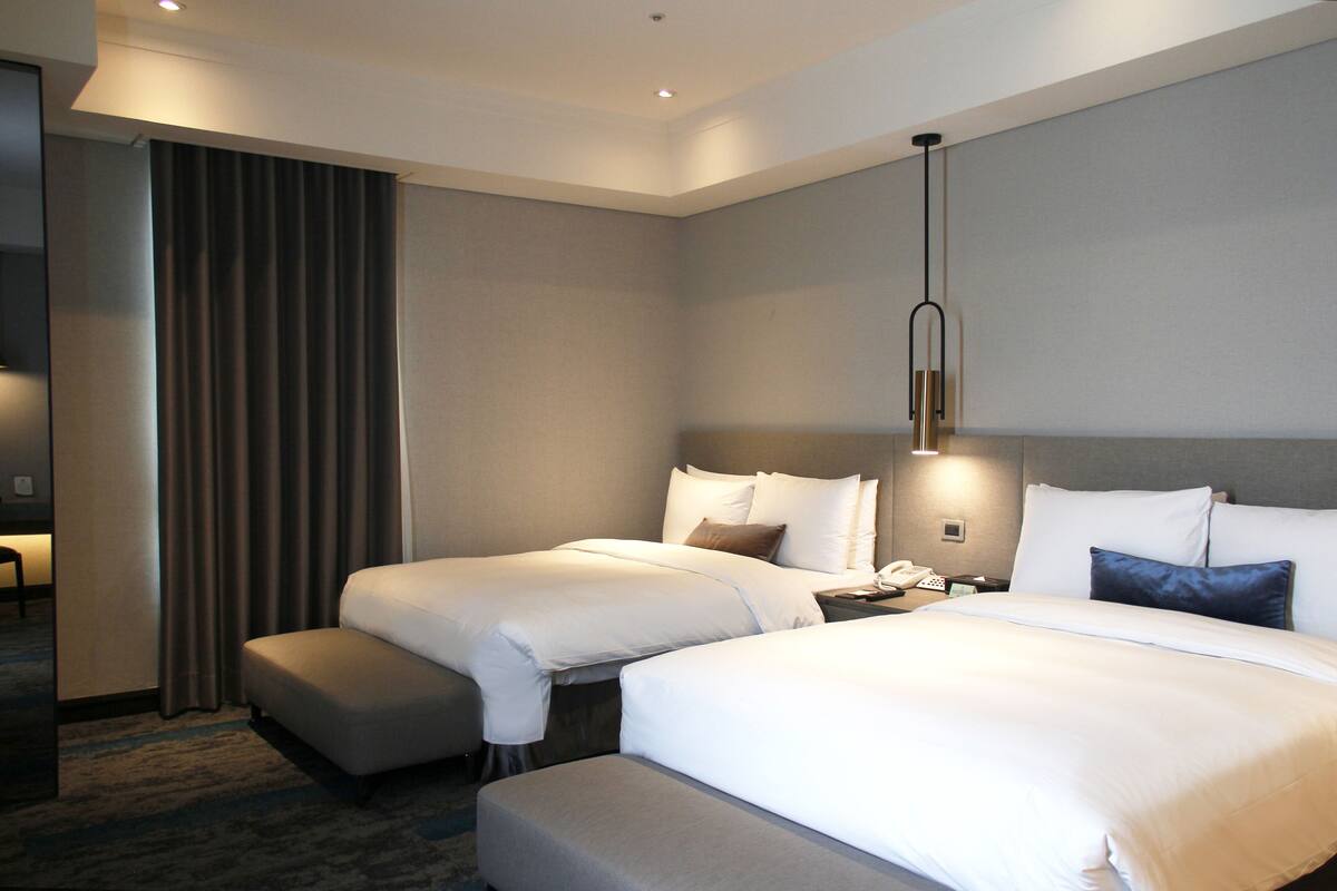 Ein Hotelzimmer mit zwei Betten, einem Schreibtisch, einem Stuhl und einem großen Fenster mit Vorhängen.