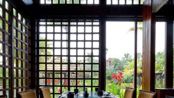 Une table à manger dressée avec les ustensiles traditionnels d’une cérémonie du thé chinoise, entourée de chaises en bois, avec vue sur la verdure et une fenêtre ornée d’un motif en treillis.