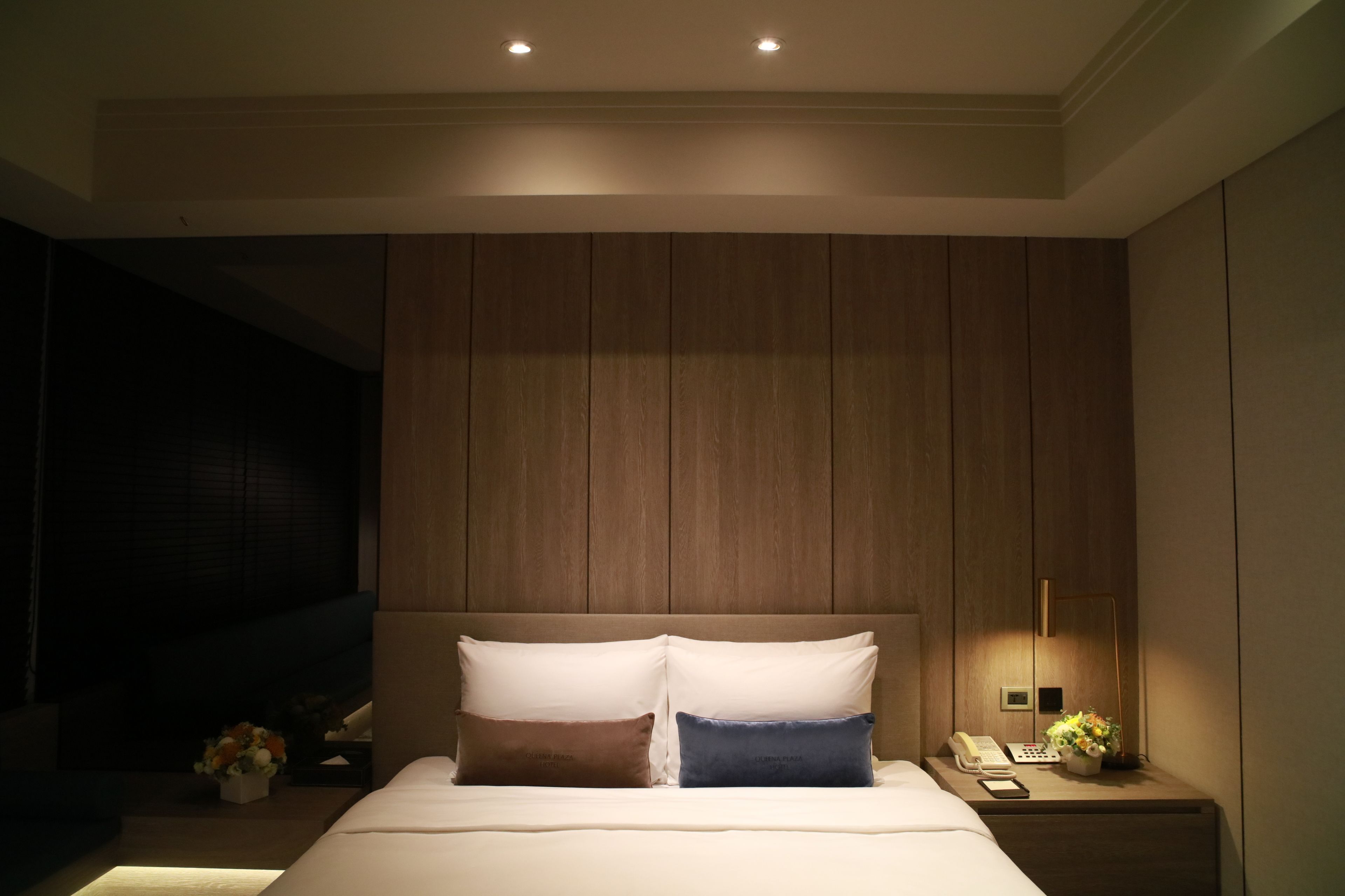 Une chambre d’hôtel moderne avec un grand lit, des murs recouverts de panneaux de bois, une table de chevet avec une lampe et une petite table de nuit avec un téléphone et des fleurs.