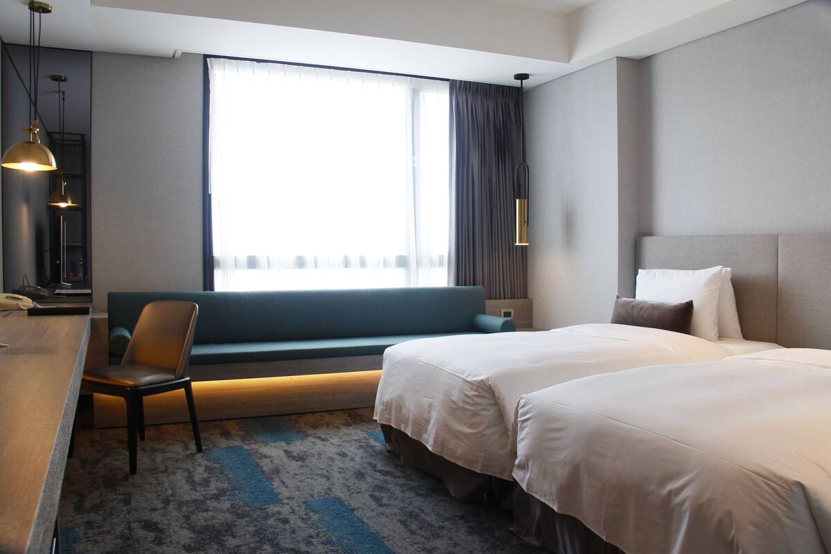 Ein Hotelzimmer mit einem Bett, einem Schreibtisch, einem Stuhl, einer Couch und einem großen Fenster mit Vorhängen.