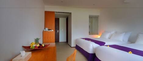 A hotel room with two beds, a wooden desk, a small table with a tissue box, and a bowl of fruit.