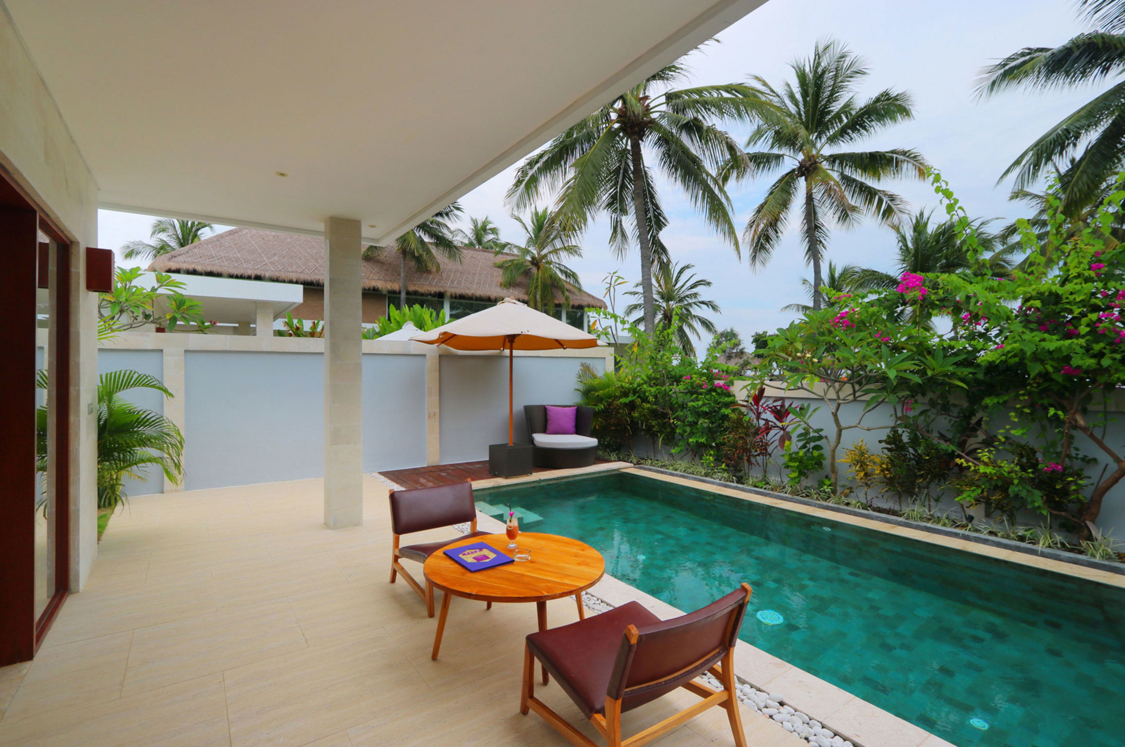 Un área junto a la piscina con mesa y sillas, una estructura de techo de paja y exuberante vegetación tropical.